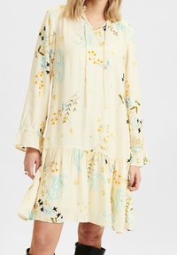 Knee-length krémové šaty s dlouhými rukávy, stahovací kapucí a květinovými vzory v modré, oranžové a zelené barvě. Volánkový spodní lem.