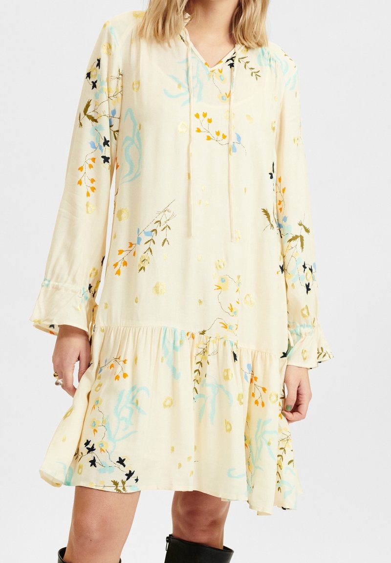 Knee-length krémové šaty s dlouhými rukávy, stahovací kapucí a květinovými vzory v modré, oranžové a zelené barvě. Volánkový spodní lem.