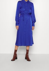 Kungsblå, långärmad klänning med knappstängning framtill, midja som är insvängd och strukturerad tyg. Kombinerad med bruna, lackläder ankelskor.