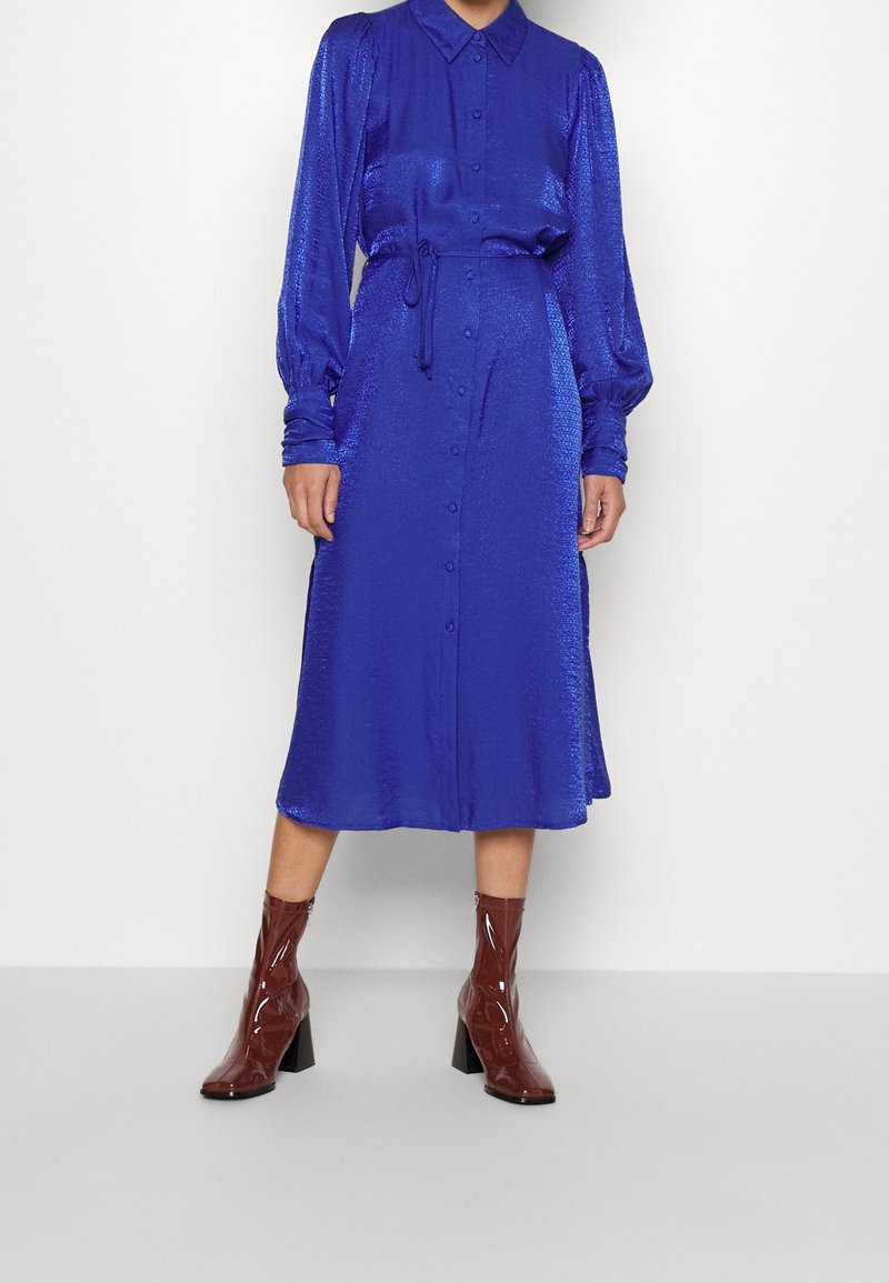 Kungsblå, långärmad klänning med knappstängning framtill, midja som är insvängd och strukturerad tyg. Kombinerad med bruna, lackläder ankelskor.