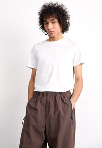 Balta medvilnės marškinėliai, trumpomis rankovėmis; rudi elastiniai kelnių juostos su užtrauktukais ir šoninėmis kišenėmis, su mažo juodo logotipo detale.