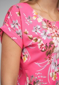 Blusa de manga corta con estampado floral en rosa intenso con un acabado texturizado. Presenta una mezcla de flores blancas, amarillas y marrones de diferentes tamaños.