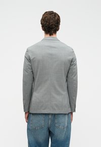 Άν�δρας με σγουρά μαλλιά φορώντας ανοιχτό γκρι σακάκι και μπλε τζιν, στέκοντας με την πλάτη γυρισμένη σε έναν απλό λευκό φόντο.