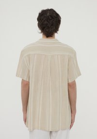 Camicia a maniche corte di un leggero beige con righe verticali. Realizzata in un tessuto texturizzato, presenta un collo posteriore e una vestibilità morbida.