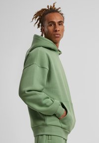 Sweatshirt à capuche en tissu vert doux, avec une poche kangourou, des bords côtes et une coupe décontractée. Texture lisse, design minimal.