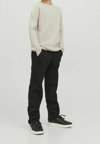 Barn står med händerna i fickorna iförda en beige tr�öja, svarta byxor och svarta sneakers med vita sulor mot en enkel bakgrund.