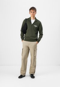 Olivengrøn zip-up sweater med rib strik ved håndleddene, parret med beige cargobukser med flere lommer og sorte sneakers. Sort crossover taske.