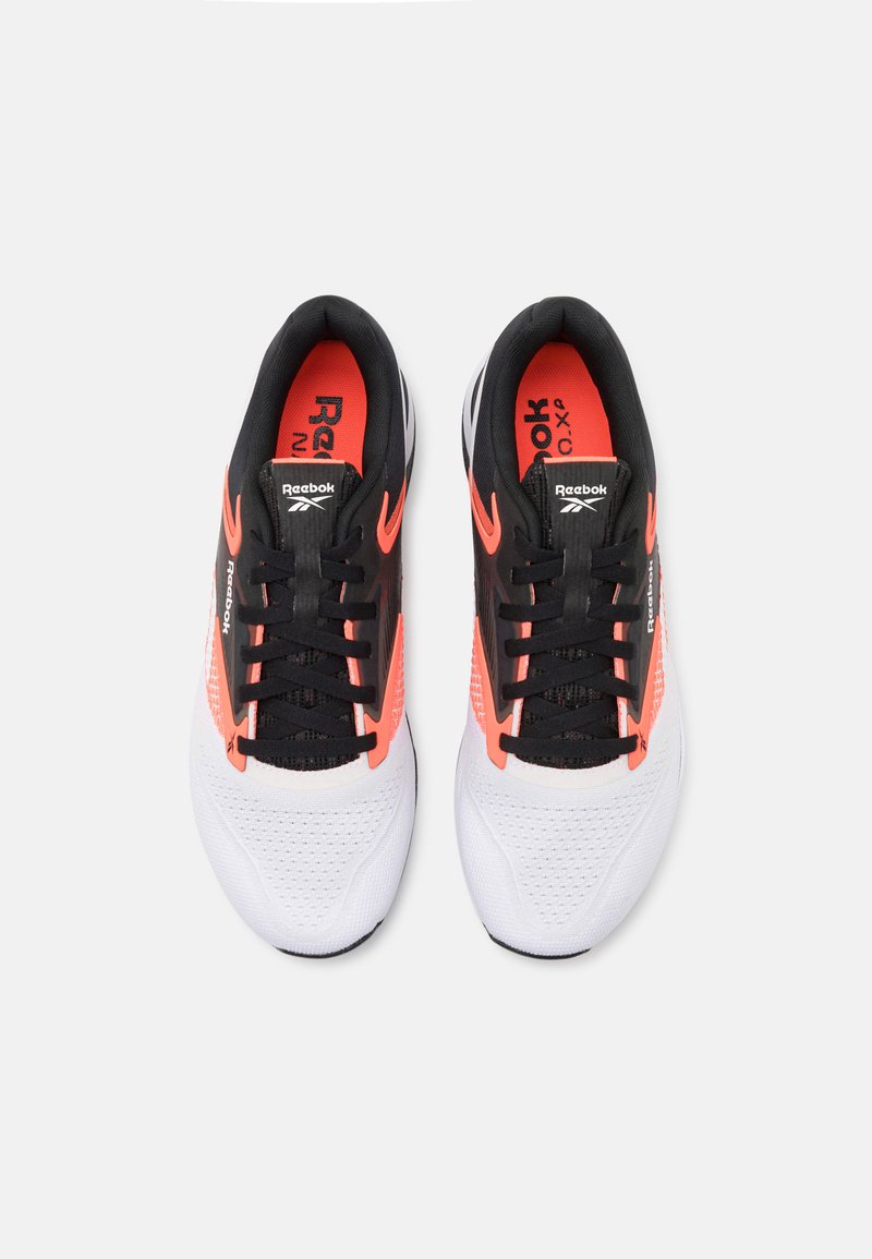 Zapatillas deportivas en blanco y negro con acentos naranjas, con parte superior de malla, punta redondeada y suela texturizada. Los cordones son negros y la marca es visible.