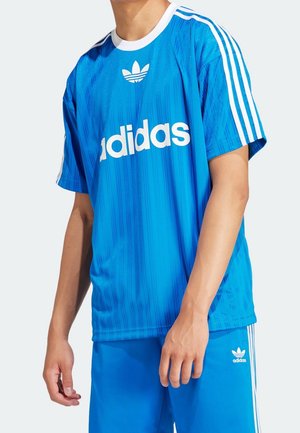 Persoon die een blauw Adidas sportoutfit draagt met witte logo's en strepen op de mouwen en de korte broek, staand tegen een effen achtergrond.