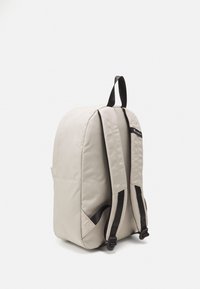 Beige backpack with a smooth fabric texture, padded shoulder straps, and a top handle. Features black accents and a subtle logo.