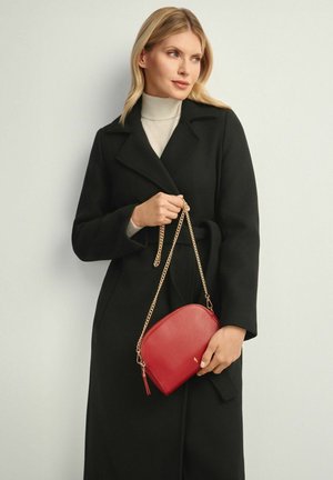 Czarny wełniany płaszcz o dopasowanym kroju, w połączeniu z czerwoną skórzaną torebką na ramię z złotym łańcuszkiem i gładką fakturą.