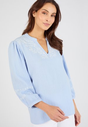 Mujer con blusa azul claro texturizada con bordado floral blanco en el escote y las mangas, posando frente a un fondo liso.