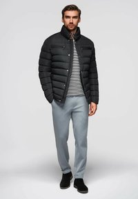 Černá péřová bunda s vysokým límcem, zipovými kapsami, pruhovanou košilí pod ní, světle šedými kalhotami a černými kotníkovými botami. Jednoduché pozadí.