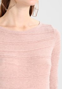 Rosa Strickpullover mit horizontalen strukturierten Streifen, aus weichem Material und rundem Ausschnitt. Große geometrische Metallohrringe runden den Look ab.
