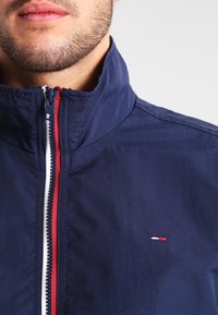 Mörkblå jacka med hög krage, utrustad med en kontrasterande röd dragkedja och en liten logotyp på bröstet. Slät textur, avslappnad design.