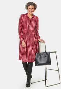 Abito rosso con motivi geometrici, maniche lunghe, colletto e vita stretta. Borsa a spalla nera in pelle e stivali neri al ginocchio. Sedia semplice.
