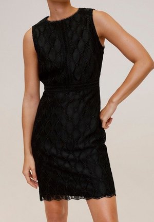 Robe noire sans manches en dentelle avec motifs floraux, taille cintrée et ourlet festonné portée par une personne debout, une main sur la hanche.