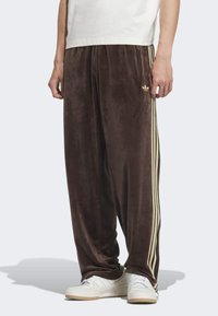 Hnedé velúrové tepláky so zlatými bočnými pruhmi a malými logami. Voľný strih s jemnou textúrou, kombinované s bielymi teniskami.