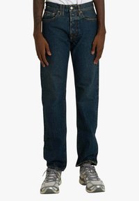 Jeans in denim blu scuro con vestibilità a gamba dritta, chiusura con bottoni e design standard a cinque tasche. Indossati con scarpe da ginnastica grigie.