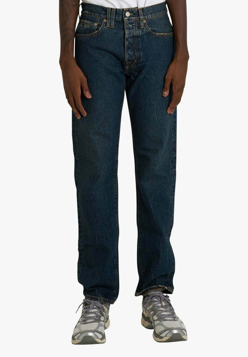 Jeans in denim blu scuro con vestibilità a gamba dritta, chiusura con bottoni e design standard a cinque tasche. Indossati con scarpe da ginnastica grigie.