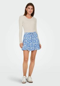 Licht beige geribbelde top met lange mouwen, gecombineerd met een blauwe rok met bloemenpatroon. De rok heeft een gelaagd ontwerp en een gerimpelde zoom. Witte sneakers zijn inbegrepen.