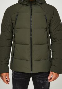 Mand iført en olivengrøn polstret jakke med sorte lynlåslommer og mørke jeans, står mod en lys baggrund.