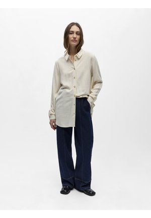 Žena stojící v uvolněné béžové košili na knoflíky, širokých tmavě modrých kalhotách a černých botách na jednorodém světlém pozadí.