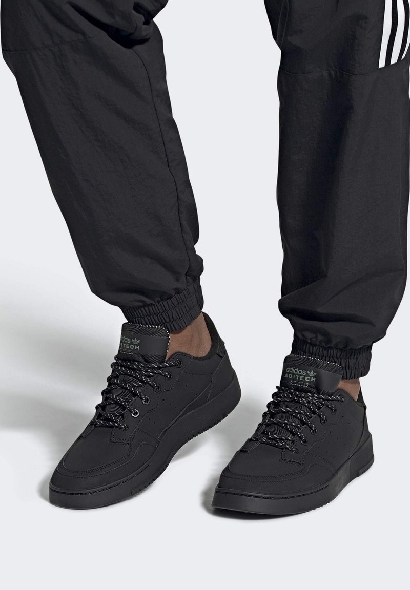 Scarpe sportive nere in materiale sintetico liscio, con un design a bassa regolazione, lacci testurizzati e logo sulla linguetta e sul lato.