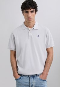 Polo de color gris claro con cuello clásico, tapeta de tres botones y un logo de calavera en navy en el pecho. Mangas cortas y tejido de algodón.