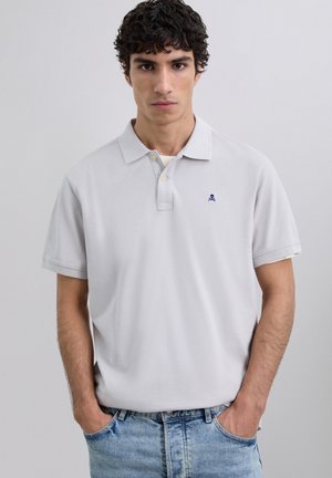 Polo de color gris claro con cuello clásico, tapeta de tres botones y un logo de calavera en navy en el pecho. Mangas cortas y tejido de algodón.