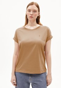 Braunes kurzärmliges T-Shirt mit Rundhalsausschnitt, aus weichem Material gefertigt. Mit umgeschlagenen Ärmeln und kleinem Logo auf der Brust. Kombiniert mit blauen Jeans.