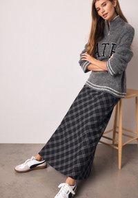 Pull gris en maille avec une fermeture éclair, décoré d'un grand texte noir. En dessous, une longue jupe maxi à carreaux en noir et gris. Des baskets blanches complètent le look.