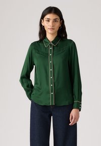 Grüne Satinbluse mit kragenförmigem Ausschnitt, vorderem Knopfverschluss und kontrastierendem goldenen Besatz. Verfügt über zwei Brusttaschen und lange Ärmel.