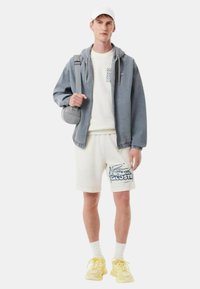 Sudadera gris con cremallera, sudadera blanca, pantalones cortos crema con logo azul, zapatillas amarillas y un bolso cruzado gris. Diseño limpio, estilo casual.