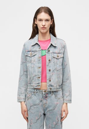 Moteris vilki šviesiai mėlyną džinsinę striukę ir džinsus su rožinius gėlių raštais, derinama su ryškiai rožiniu trumpu viršumi, fone paprasta.