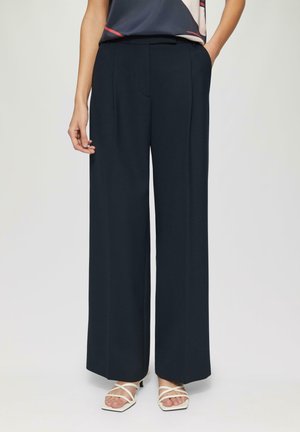 Pantalon large en tissu bleu marine foncé, doté d'une taille haute, de plis avant et de poches latérales. Associé à des sandales blanches à lanières.