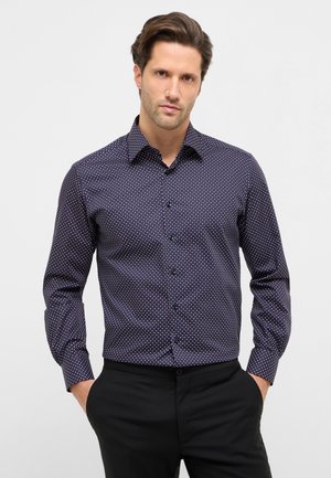 Mørkeblå skjorte med hvide og røde polkaprikker, lange ærmer, knappes foran og klassisk krave. Stoffet ser glat og tætsiddende ud.