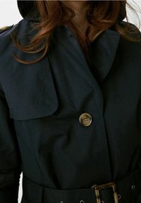 Trench-coat imperméable noir avec un grand col, fermeture à boutons dorés et taille ceinturée. Présente des détails cousus et une texture lisse.