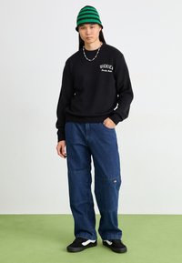 Maglione nero con logo "Dickies", pantaloni in denim blu a gamba larga, sneakers nere, cappello a maglia a righe verdi e nere, collana di perline. In piedi su un pavimento verde.