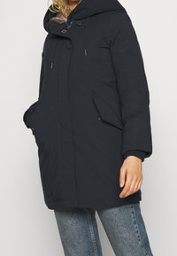 Tummansininen vuorattu takki, jossa on turkiskauluksen huppu, kaksi sivutaskua, nepparit ja resorit hihansuissa. Pehmeä, teksturoitu kangas.