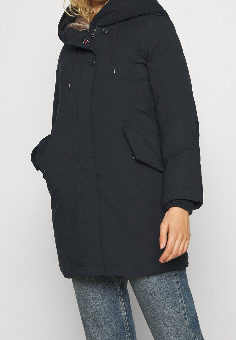 Tummansininen vuorattu takki, jossa on turkiskauluksen huppu, kaksi sivutaskua, nepparit ja resorit hihansuissa. Pehmeä, teksturoitu kangas.