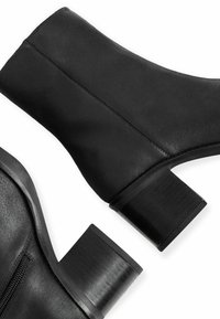 Par de botines de cuero negro con tacones de bloque y cremallera lateral, mostrados desde ángulos lateral y trasero sobre fondo blanco.