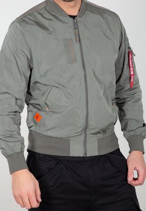 Homme portant une veste bomber grise avec une fermeture éclair frontale, une étiquette orange près de la poche, et une étiquette rouge "Remove Before Flight" sur la manche gauche.
