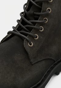 Botín de ante negro con punta redondeada, ojales metálicos y suela gruesa negra con detalles texturizados y cordones del mismo color.