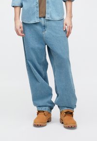 Vaqueros de mezclilla azul claro con un corte holgado, combinados con botas de trabajo color marrón claro. La camisa es de un azul claro, con botones y ligeramente holgada.