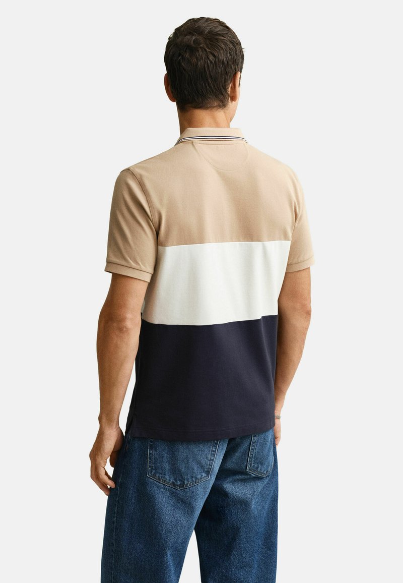 Homme portant un polo à manches courtes avec des blocs de couleur beige, blanc et marine, associé à un jean bleu, vu de dos.