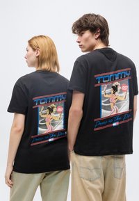 Δύο νέοι ενήλικες φορούν μαύρα γραφικά t-shirts με ρετρό σχεδιασμό γυμναστικής και κείμενα "Tommy" και "Pump up the Pop" στην πλάτη.