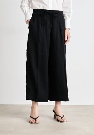 Pantaloni neri a gamba larga con vita regolabile tramite cordoncino, dotati di tasche laterali e una texture liscia, abbinati a décolleté nere con cinturini.