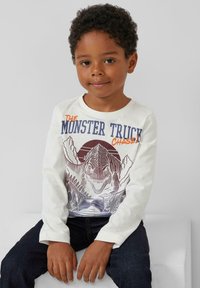 Langärmliges weißes Hemd mit Dinosauriergrafik und Bergdesign in Grau und Orange. Dunkelblaue Jeans im strukturierten Stil.