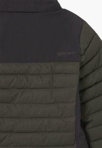 Veste isolée noire et vert foncé avec une texture matelassée, présentant un détail de logo et un tissu lisse au niveau des épaules.